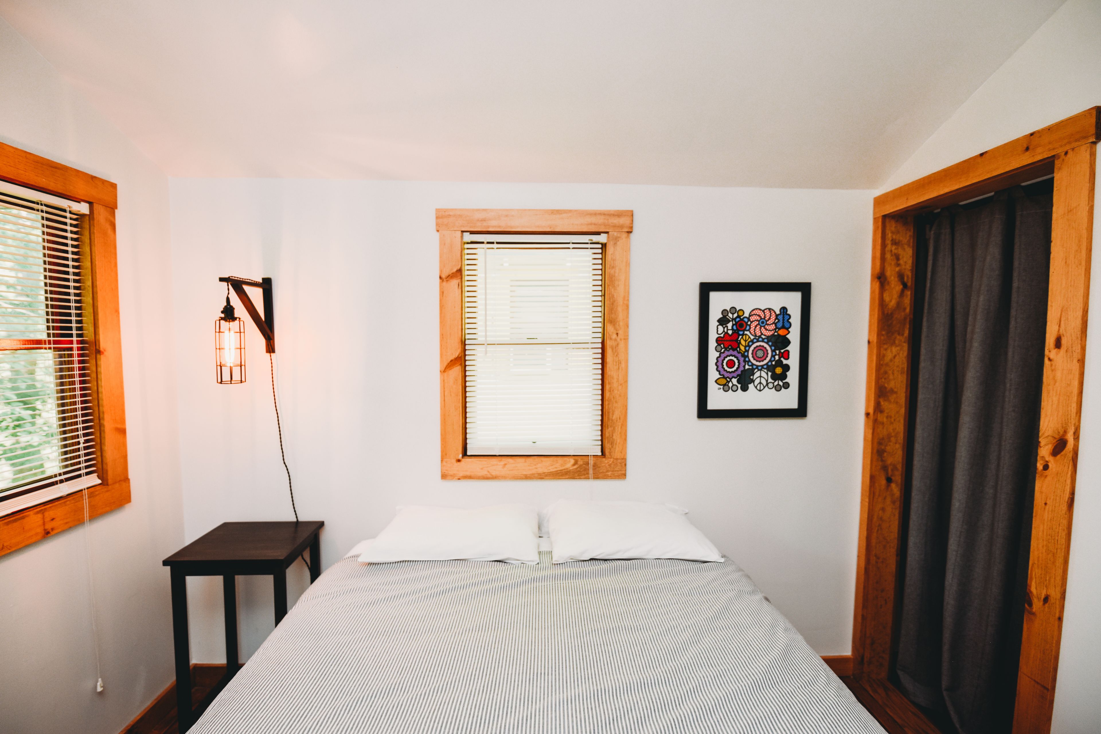 Een slaapkamer met een bed, een nachtkastje, een wandlamp, een raam met jaloezieën en een lijst met een afbeelding aan de muur.