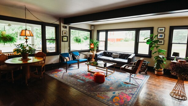 Amplia sala de estar con piso de madera, una mesa redonda con sillas, un sofá y una mesa de centro. Grandes ventanas ofrecen luz natural y vistas al exterior.