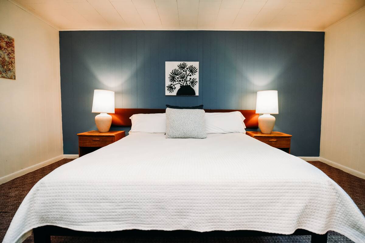 A neatly made bed with white bedding, two bedside tables with lamps, a decorative wall piece, and a dark blue accent wall.