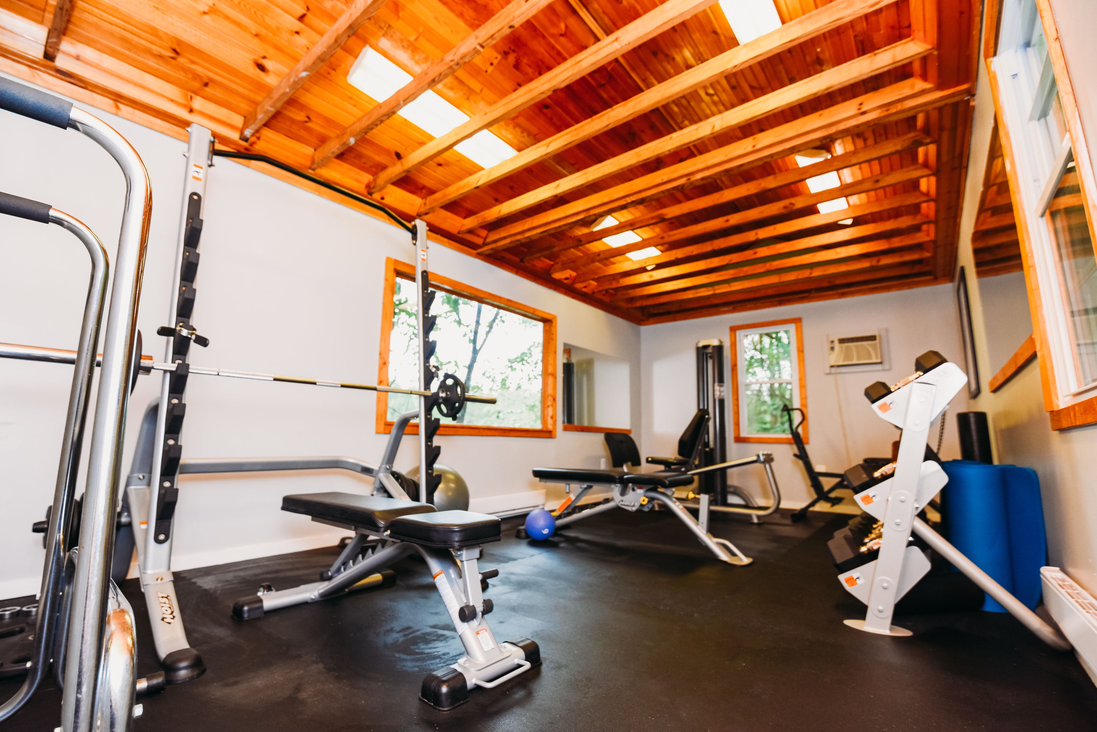 Un gimnasio con diverso equipo de ejercicio, vigas de madera en el techo y grandes ventanales.
