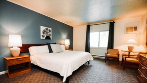 Habitación de hotel con una cama grande, dos mesitas de noche con lámparas, una silla, un aparador, una ventana con cortinas y una obra de arte en la pared.