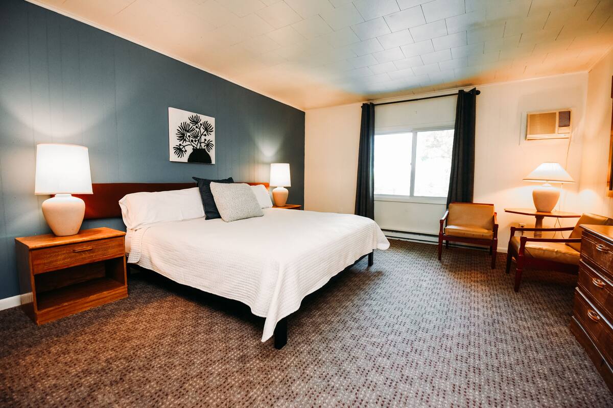 A hotel room with a large bed, two bedside tables with lamps, a chair, a dresser, a window with curtains, and a wall art piece.