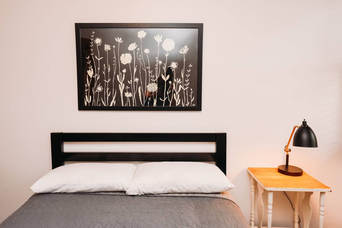 A bed with white pillows, a black headboard, and a grey quilted bedspread. Above the bed is a framed black and white floral artwork. To the right is a wooden nightstand with a black desk lamp.