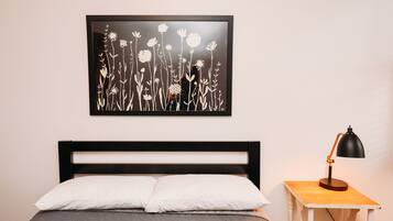 Una cama con almohadas blancas, cabecera negra y colcha acolchada gris. Encima de la cama, una obra de arte floral en blanco y negro enmarcada. A la derecha, una mesita de noche de madera con una lámpara de escritorio negra.