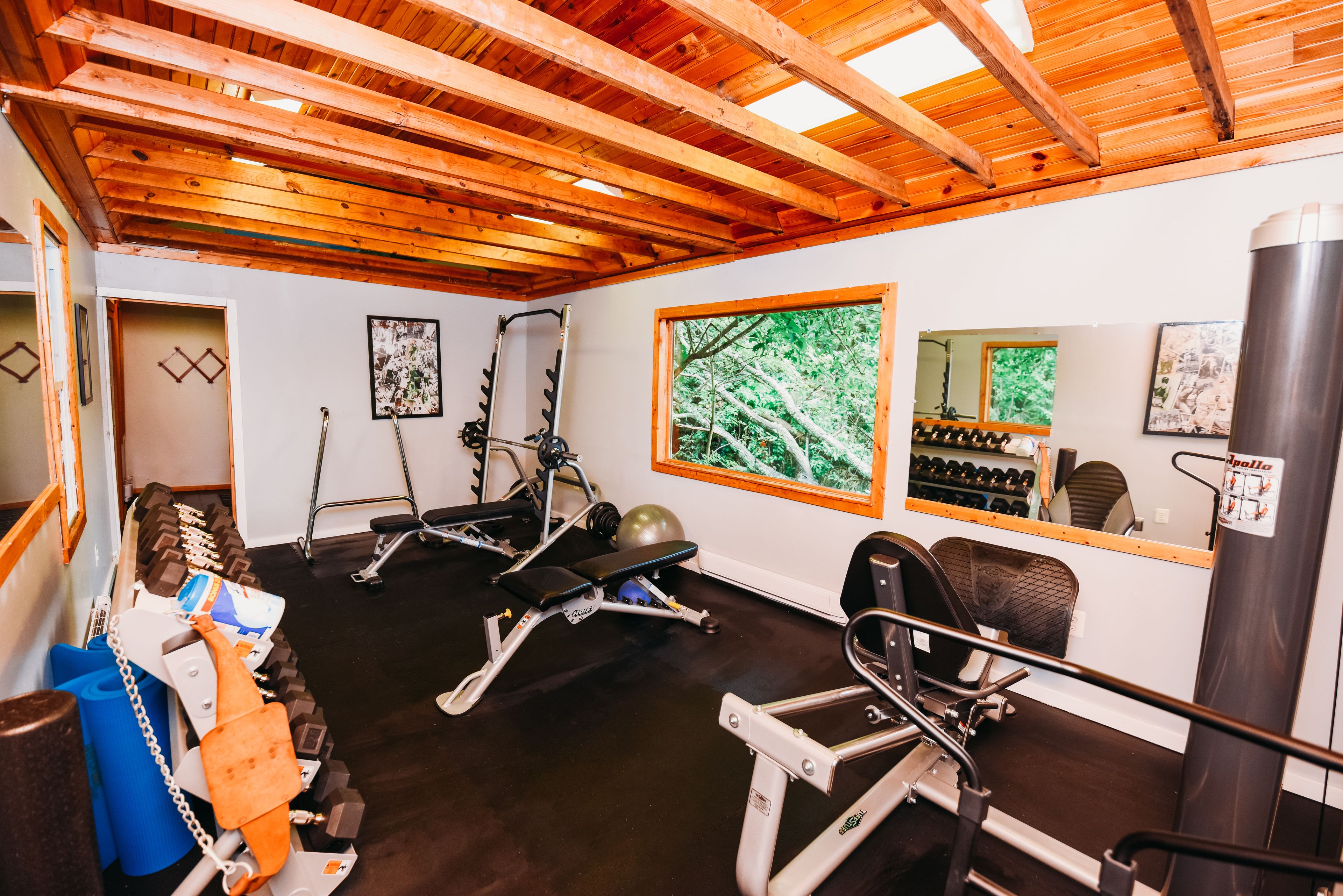 Une salle de sport équipée de matériel d’exercice, avec des poutres en bois au plafond et une grande fenêtre donnant sur des arbres.