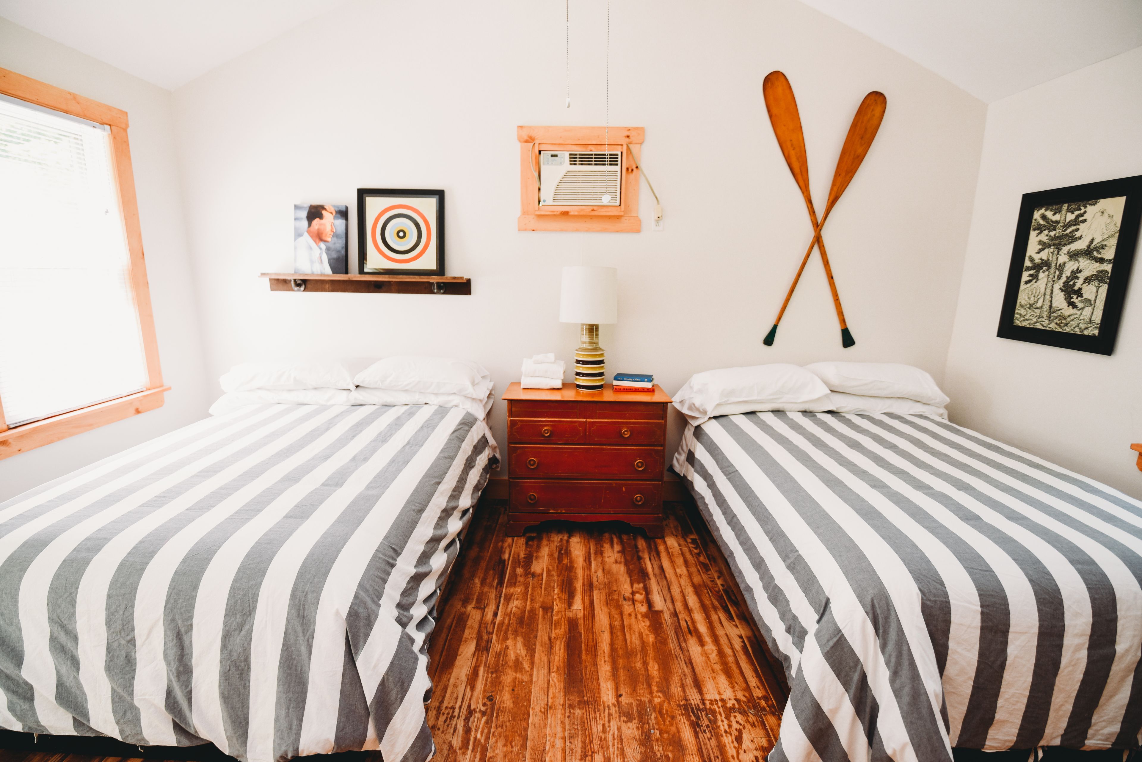 Habitación con dos camas, piso de madera, una repisa con fotografías, una lámpara y dos palas de madera en la pared.