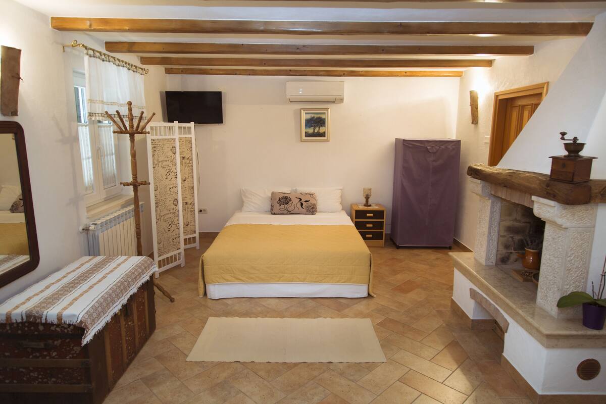 Een slaapkamer met een bed, een dressoir, een spiegel, een televisie, een open haard en een raam met gordijnen.