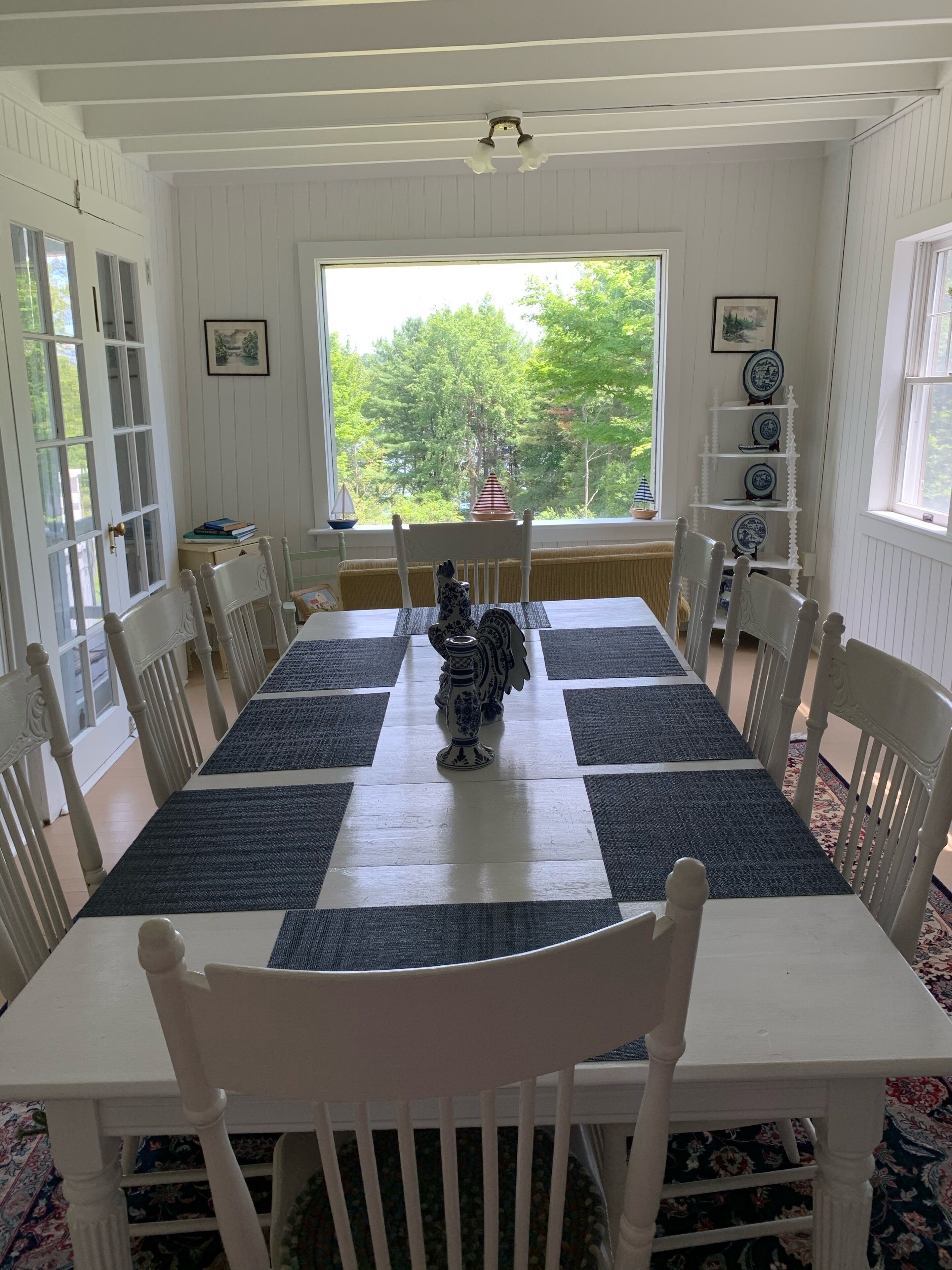 A dining room with a long table, white chairs, a decorative vase, and a view of trees through a window.