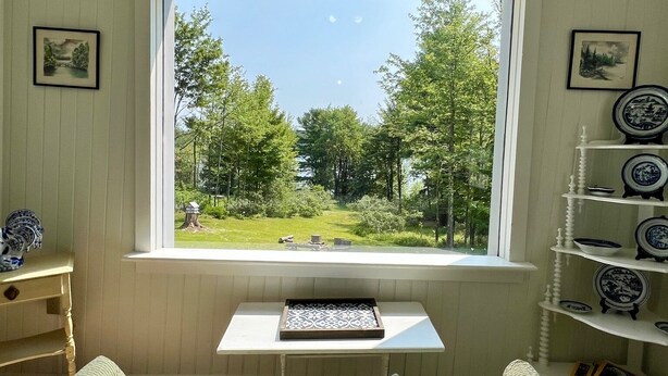 A room with a large window offering a view of a lake and trees. There is a white console table with a tray and a cushion with "The Lake" text.