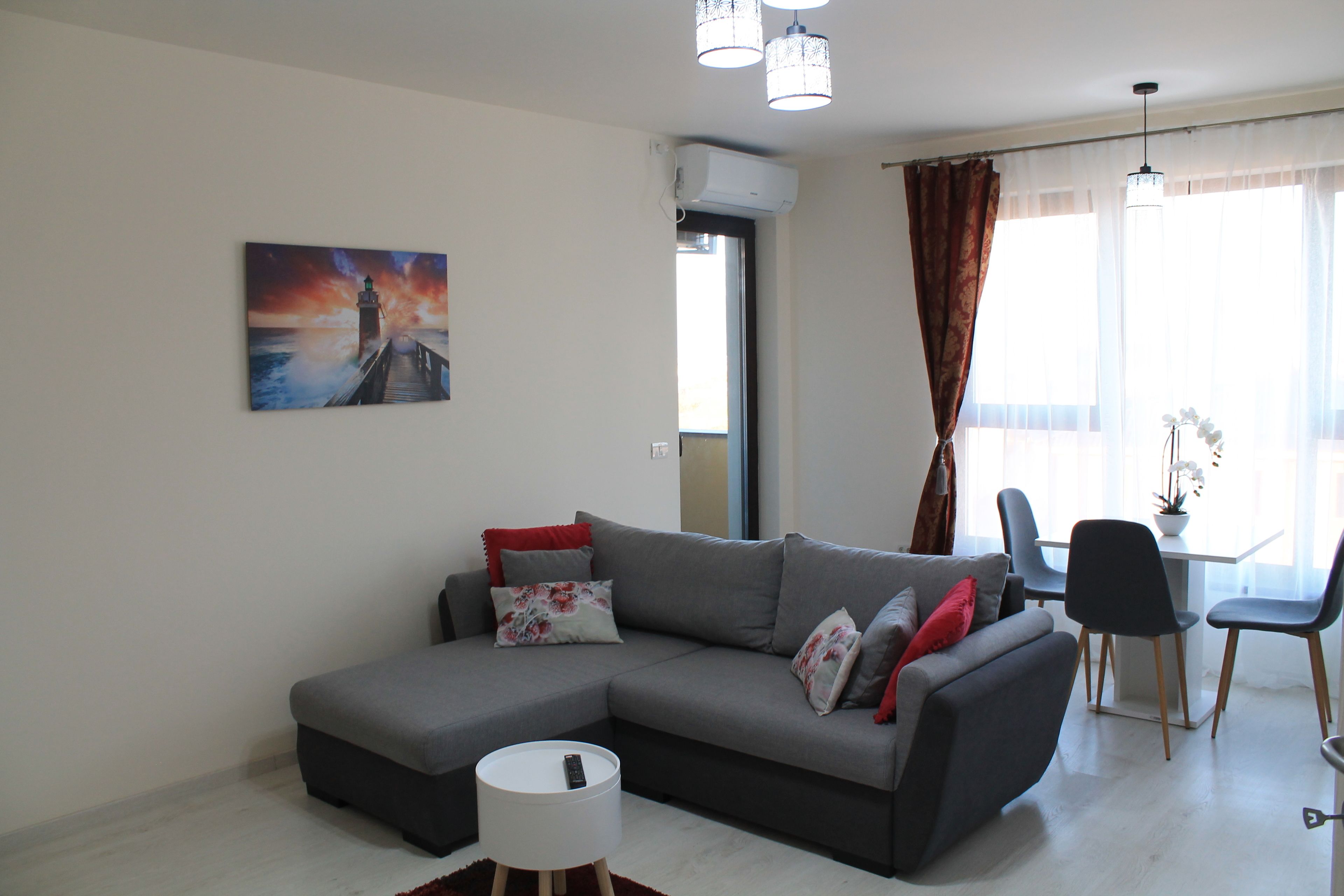 A modern living room with a grey sectional sofa, a round white side table, and a painting of a lighthouse on the wall.
