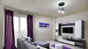 Una sala de estar moderna con un sofá gris, una mesita, un televisor montado en la pared y cortinas moradas.