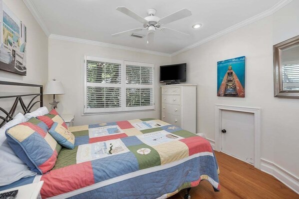 Ein Schlafzimmer mit einem Bett, einem Deckenventilator, einem Fernseher, einem Fenster mit Jalousien, einem Schminktisch und einer Tür.