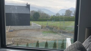 Une pièce dotée d’une grande fenêtre donnant sur un espace verdoyant, avec une clôture, un bâtiment et des montagnes à l’horizon.