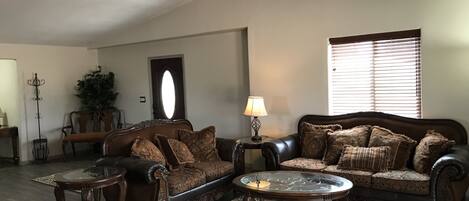 A living room with a dark brown leather sofa, a glass coffee table, and a patterned rug. There is a floor lamp and a ceiling fan.