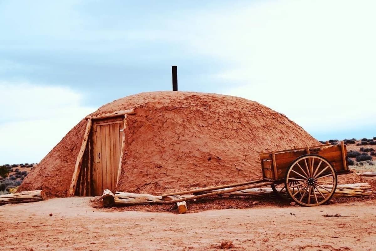 A traditional earth hut with a wooden door and a wheelbarrow in front.