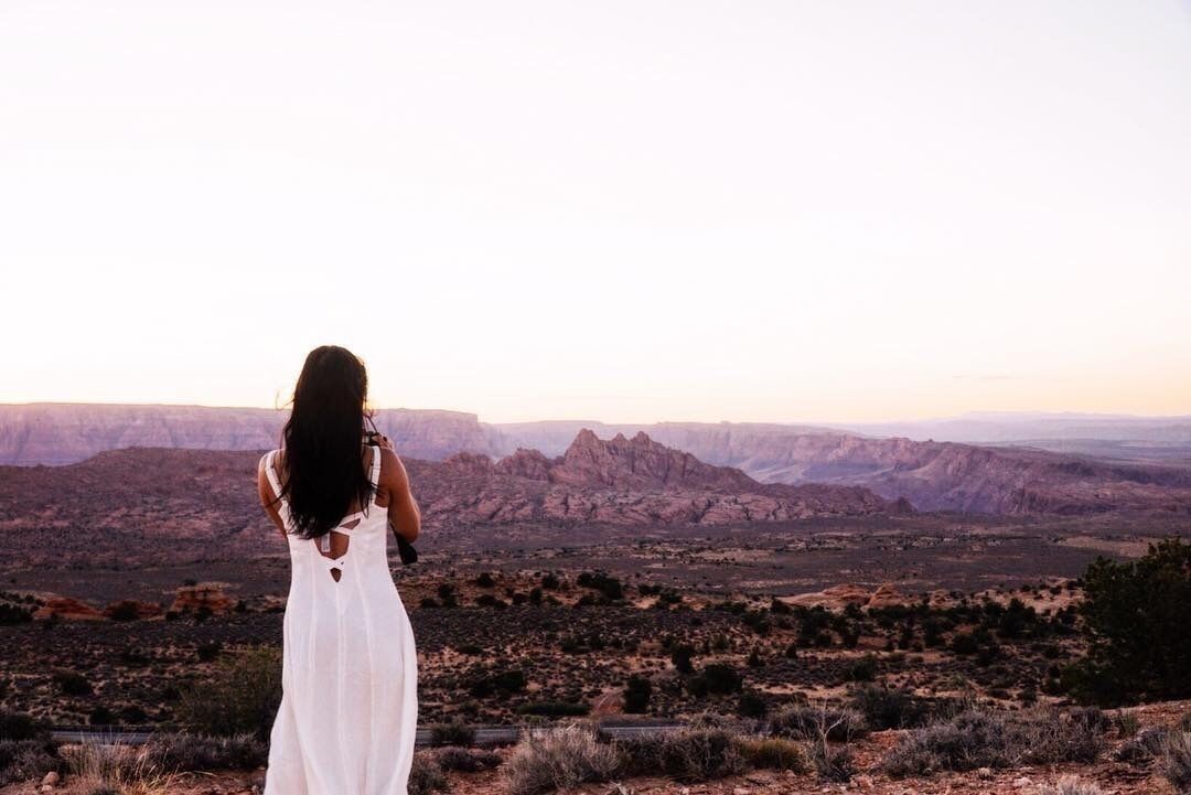A person in a white dress standing on a hill overlooking a vast canyon landscape.