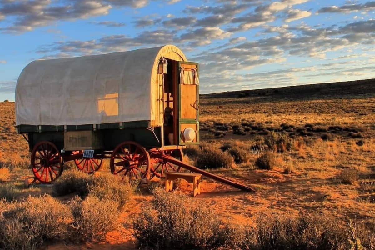 A covered wagon with a wooden door and a small table in front, set in a dry, open landscape with a clear sky.