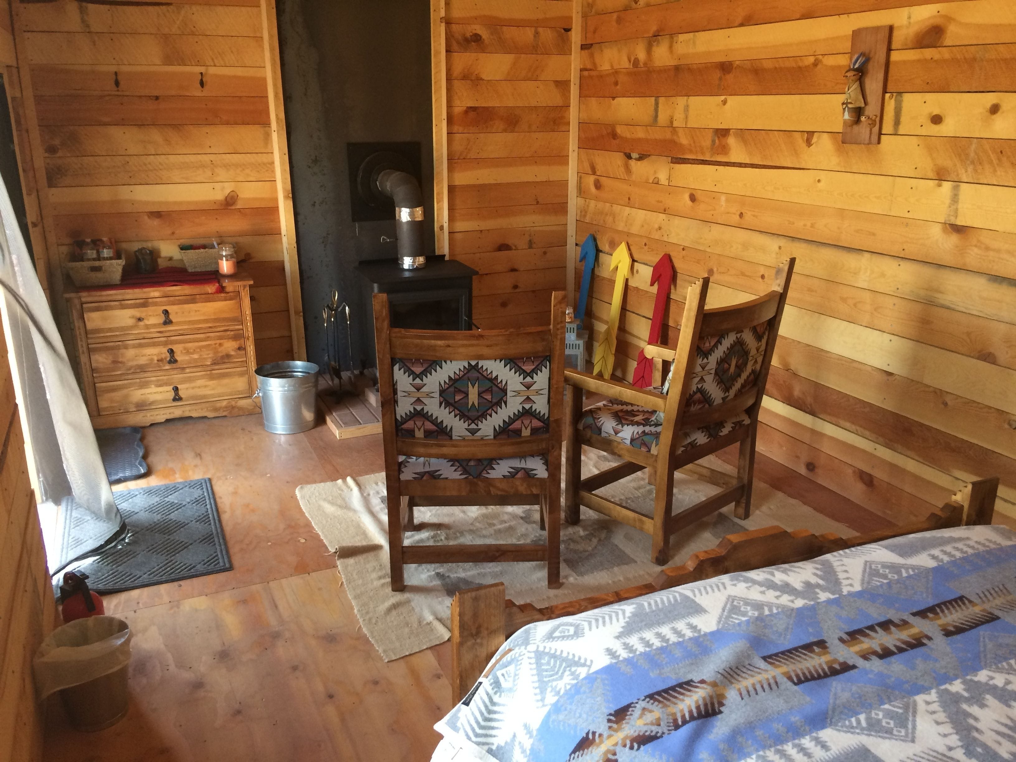 A cozy wooden cabin room with a bed, a chair with colorful cushions, a wooden dresser, and a fireplace.