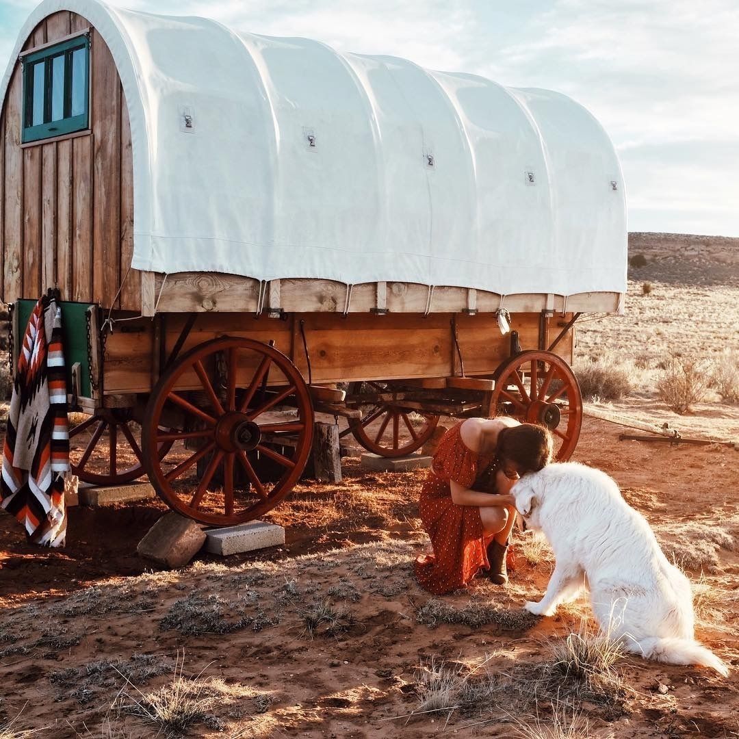 A covered wagon with wooden wheels and a white canvas roof, parked on a dirt ground with sparse vegetation.