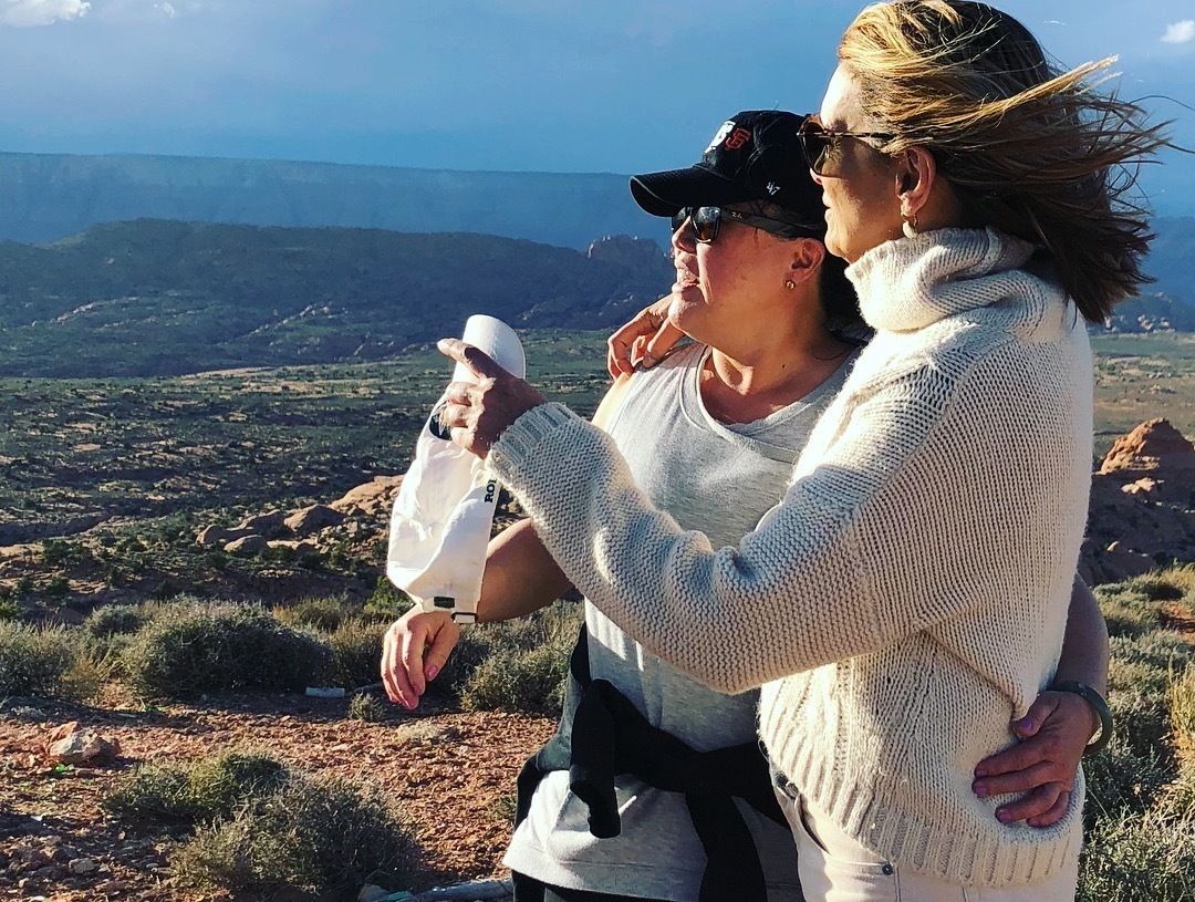 A person holding a camera, another person embracing them, and a scenic view of a mountainous landscape.