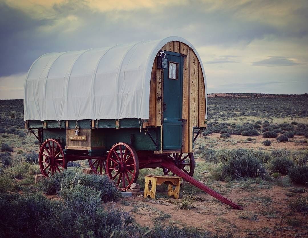 A covered wagon with a wooden structure and red wheels, situated in a desert landscape.