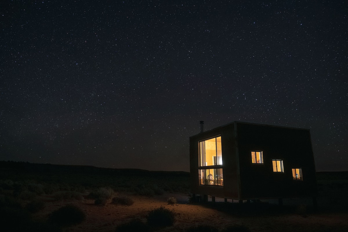 A small, cube-shaped building with large windows, situated in a desert landscape under a starry night sky.