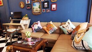 A living room with a brown sofa, a wooden coffee table, and a black and white checkered floor. There are framed pictures on the wall and a laptop on a desk. - Dluck Hostel (Kanchanaburi)
