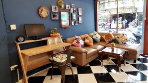 A living room with a sofa, a TV, and a coffee table. There are framed pictures on the wall and a window with a view of the street. - Dluck Hostel (Kanchanaburi)