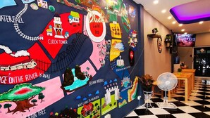 A hotel lobby with a large wall mural depicting Thailand's landmarks, a checkered floor, and a TV screen. - Dluck Hostel (Kanchanaburi)