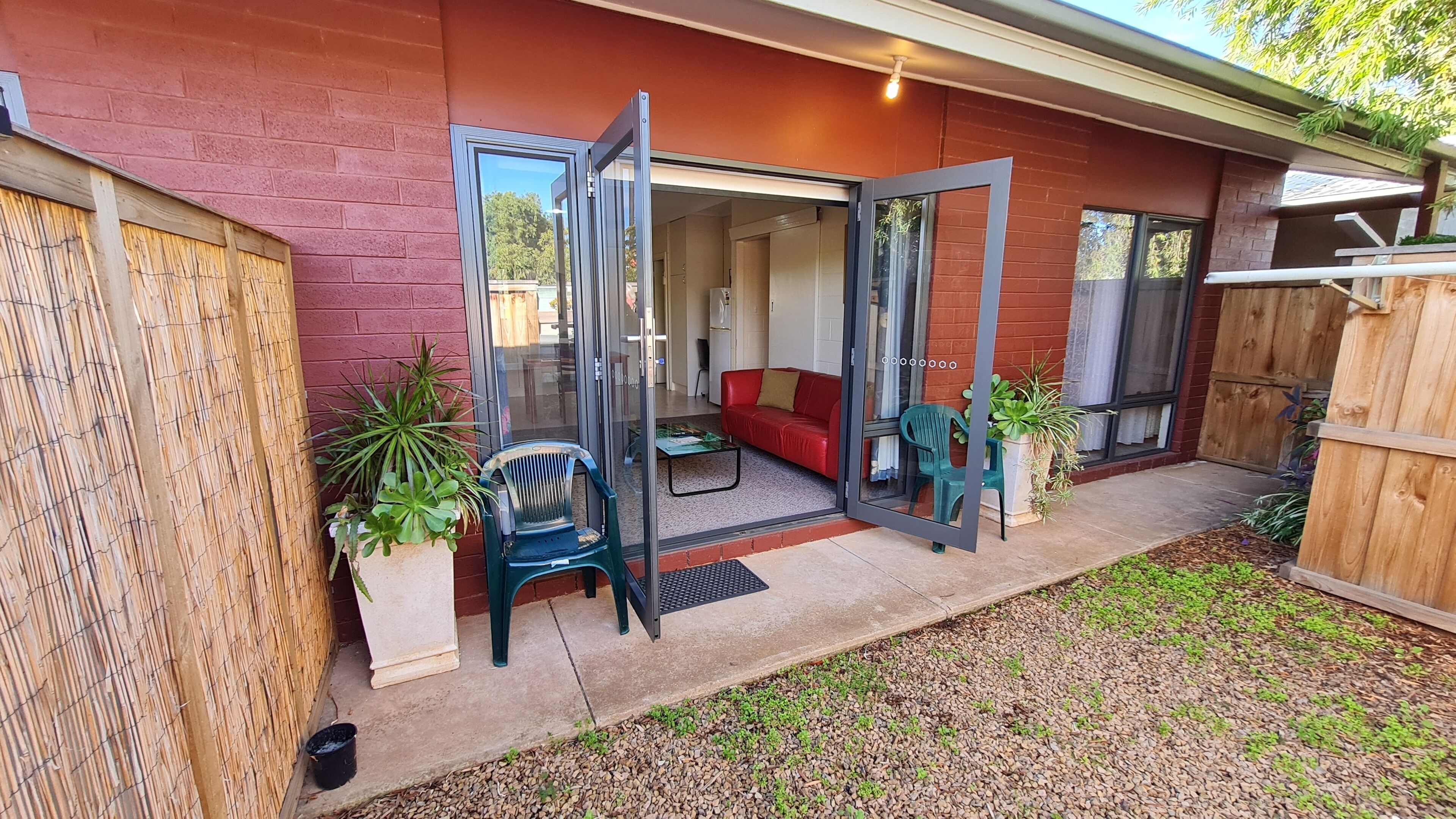 A patio area with a red brick wall, a glass door leading inside, a red sofa, and a green chair outside.