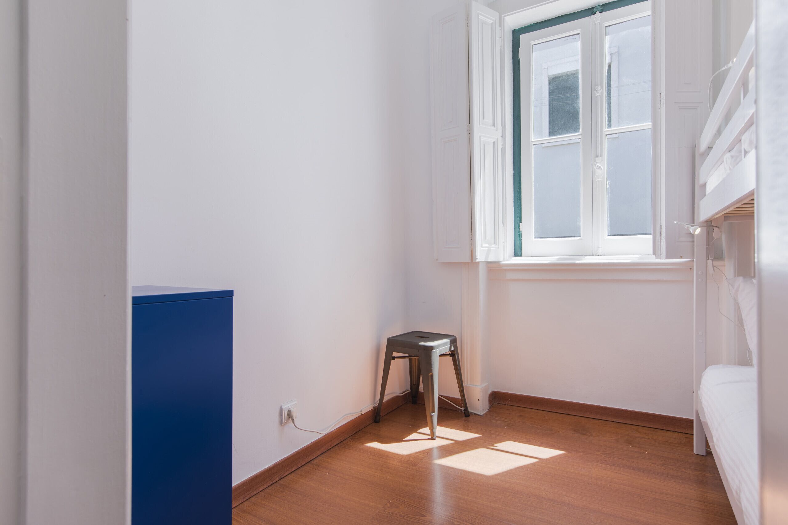 Una habitación pequeña y luminosa con suelo de madera, una ventana con persianas, una cama blanca y un armario azul.