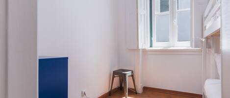 Una habitación pequeña y luminosa con suelo de madera, una ventana con persianas, una cama blanca y un armario azul.