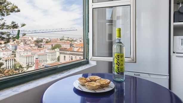 Una mesa con una fuente de quiches y una botella de vino Gazella, una ventana con vistas a edificios y un puente, y una zona de cocina con un refrigerador blanco.