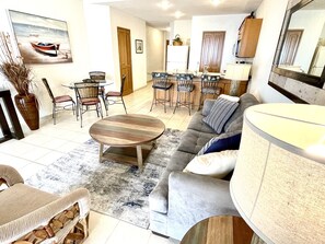 Un salon avec un canapé, une table basse et un coin repas comprenant des chaises, et une cuisine en arrière-plan.