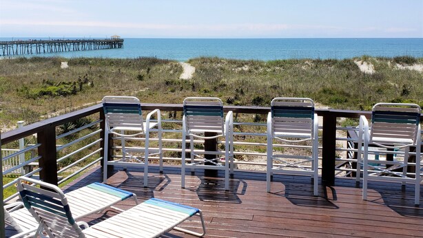 A wooden deck with metal chairs and a table overlooking a beach with a pier in the distance.