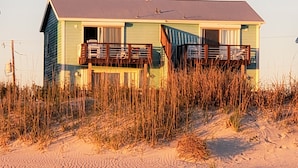 A beach house with a balcony, surrounded by sand and tall grass.