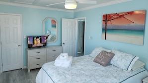 A bedroom with a bed, a ceiling fan, a painting of a beach, and a dresser with a mirror.