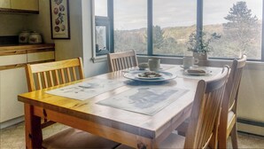 Una mesa de comedor con sillas, una ventana con vista a árboles y una zona de cocina con armarios y un cuadro en la pared.
