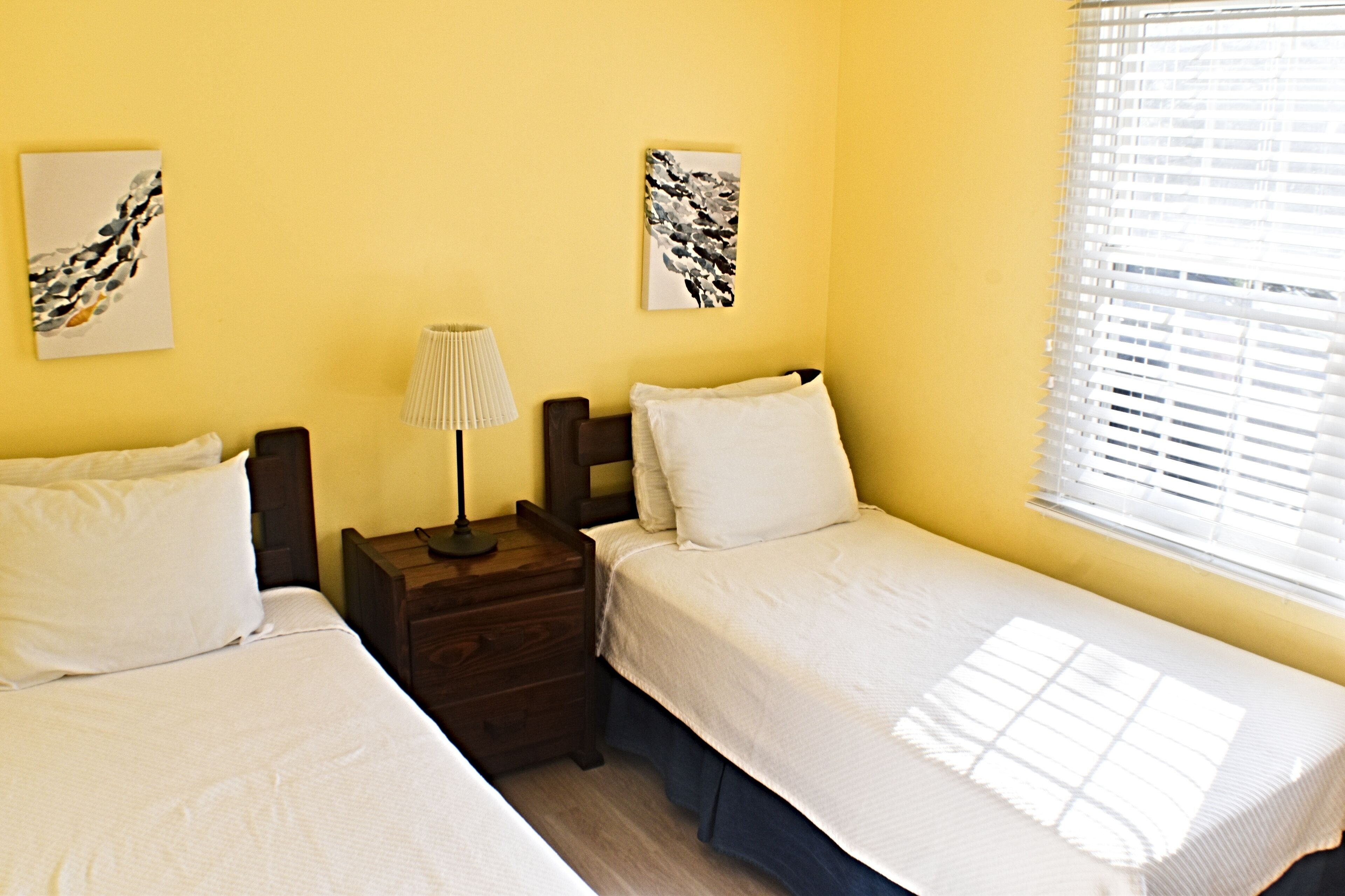 A room with two single beds, a nightstand with a lamp, and artwork on the walls.