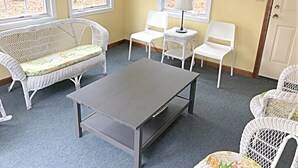 Ein Zimmer mit einem Korbsessel, einem kleinen Tisch, zwei weißen Stühlen, einer Lampe und einer Wanduhr.