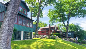 A two-story house with a green exterior and red trim, surrounded by lush greenery and trees. - Lake George Crawford House (Lake George)