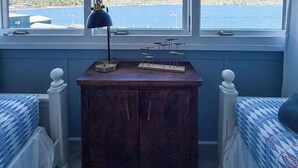 A room with two beds, a wooden nightstand with a lamp, and a window overlooking a body of water and hills. - Lake George Crawford House (Lake George)