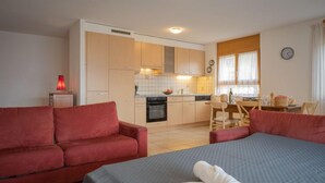 Hotellihuone, jossa on punainen sohva, sängyllä rullalle taitettu pyyhe, pieni pöytä ja taustalla näkyvä pieni keittiö.