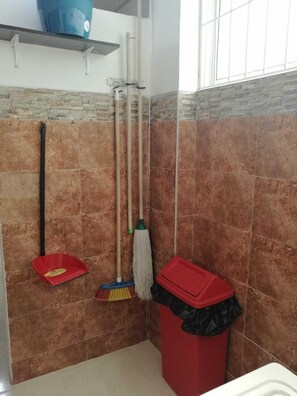 A bathroom with brown tiles, a red trash bin, and cleaning tools hanging on the wall.
