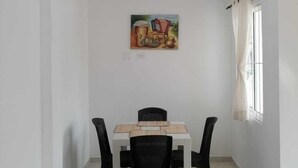 A dining area with a white table and black chairs, a painting on the wall, and a window with curtains. - Apartamentos Nayibe (Cartagena)