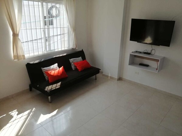 A living room with a black sofa, red and white cushions, a TV on a white stand, and a window with curtains.