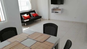 A modern living room with a dining table, two black chairs, a sofa with red and white cushions, a flat-screen TV mounted on the wall, and a small shelf below it. - Apartamentos Nayibe (Cartagena)