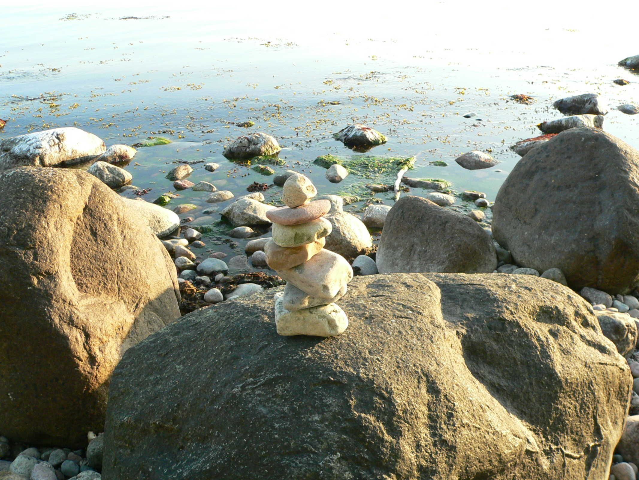 Une pile de pierres sur une plage rocheuse, avec une eau calme et des rochers épars en arrière-plan.