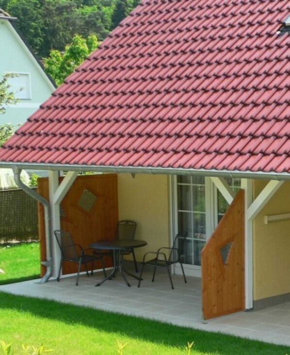 A covered patio area with a red-tiled roof, a table with chairs, and a wooden door.
