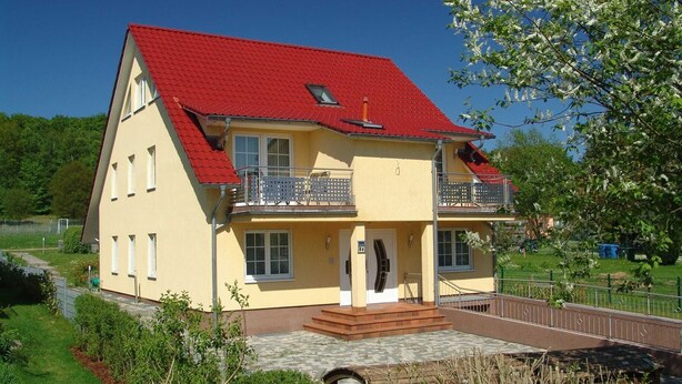 Uma casa de dois andares com telhado vermelho, uma varanda e um pequeno jardim.